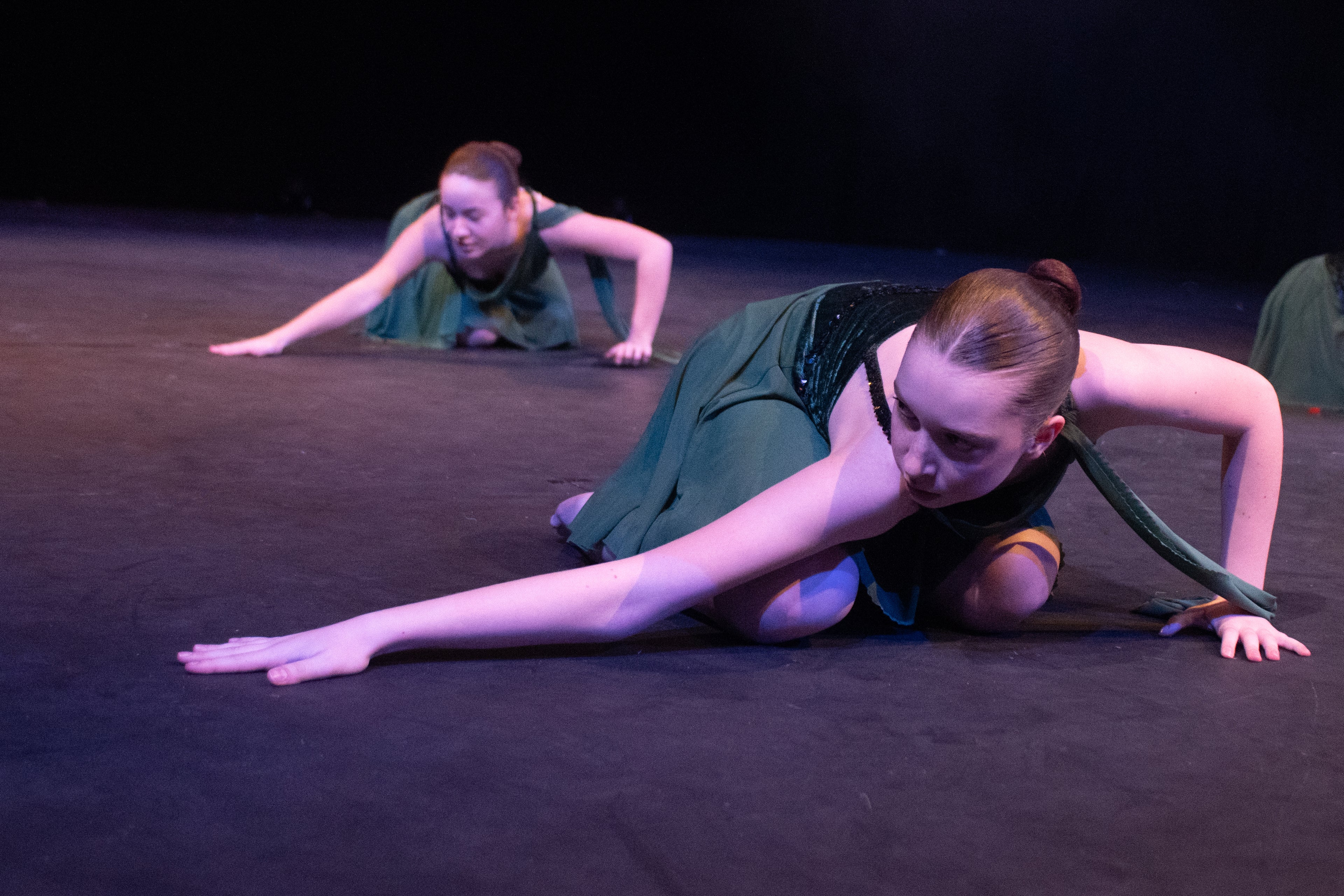 Two dancers in green outfits perform a floor routine on a dimly lit stage. They stretch forward with focus and grace, conveying intensity.