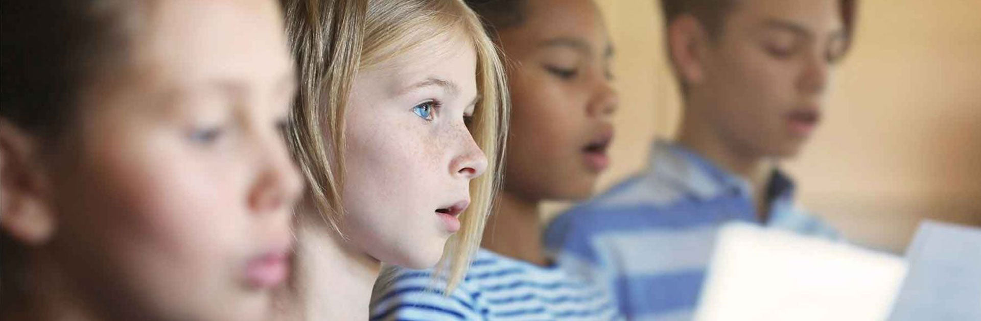 Children are side-by-side, focused and singing, holding sheets of music. They wear striped shirts, conveying a sense of concentration and harmony.