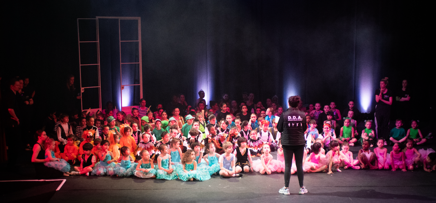 A group of young dancers in colorful costumes sit on a dimly lit stage, facing an instructor. The mood is vibrant and anticipatory, with a theatrical setting.