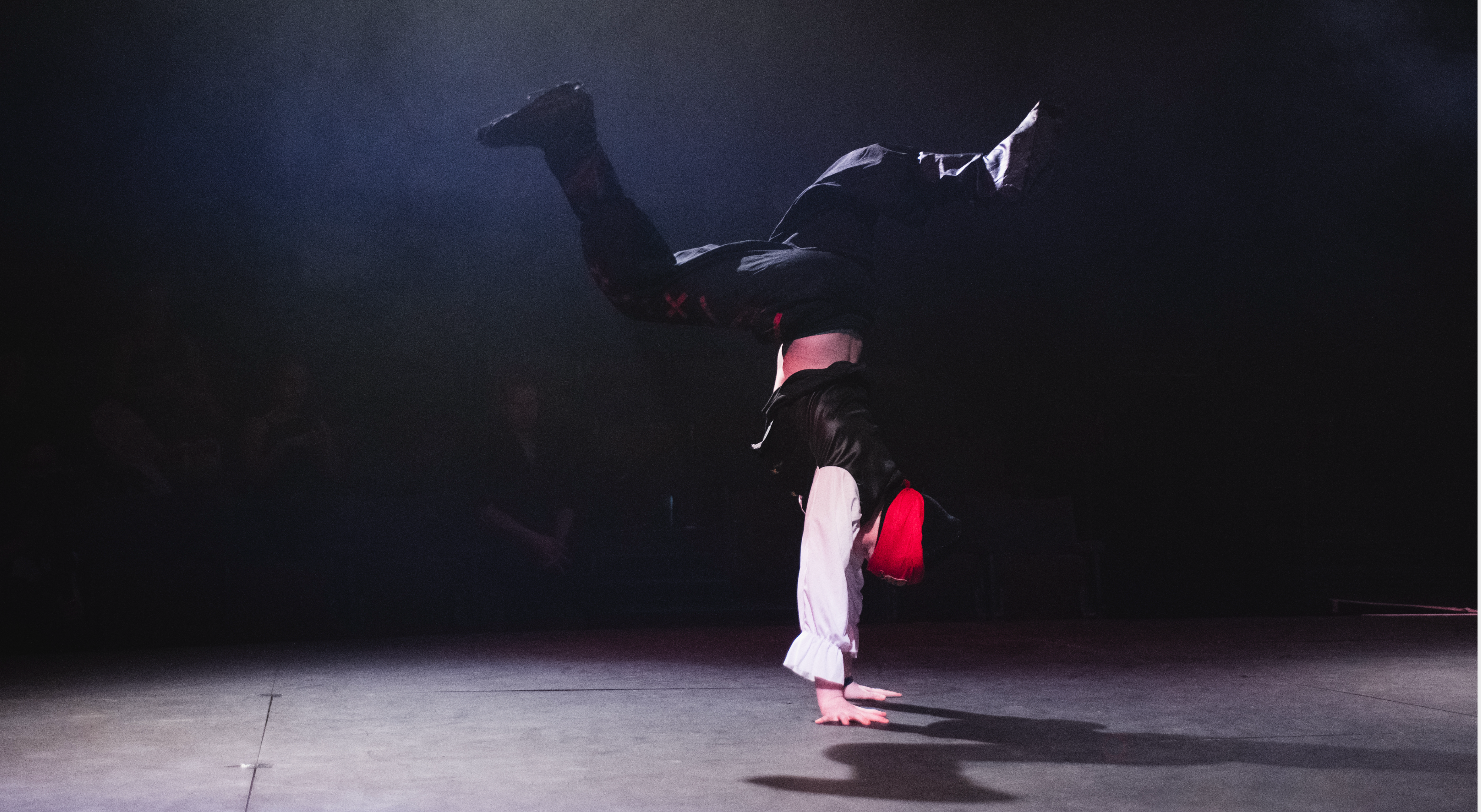 A performer in a red hat and dark costume is balancing on one hand in a dimly lit space, creating a dynamic and dramatic atmosphere.