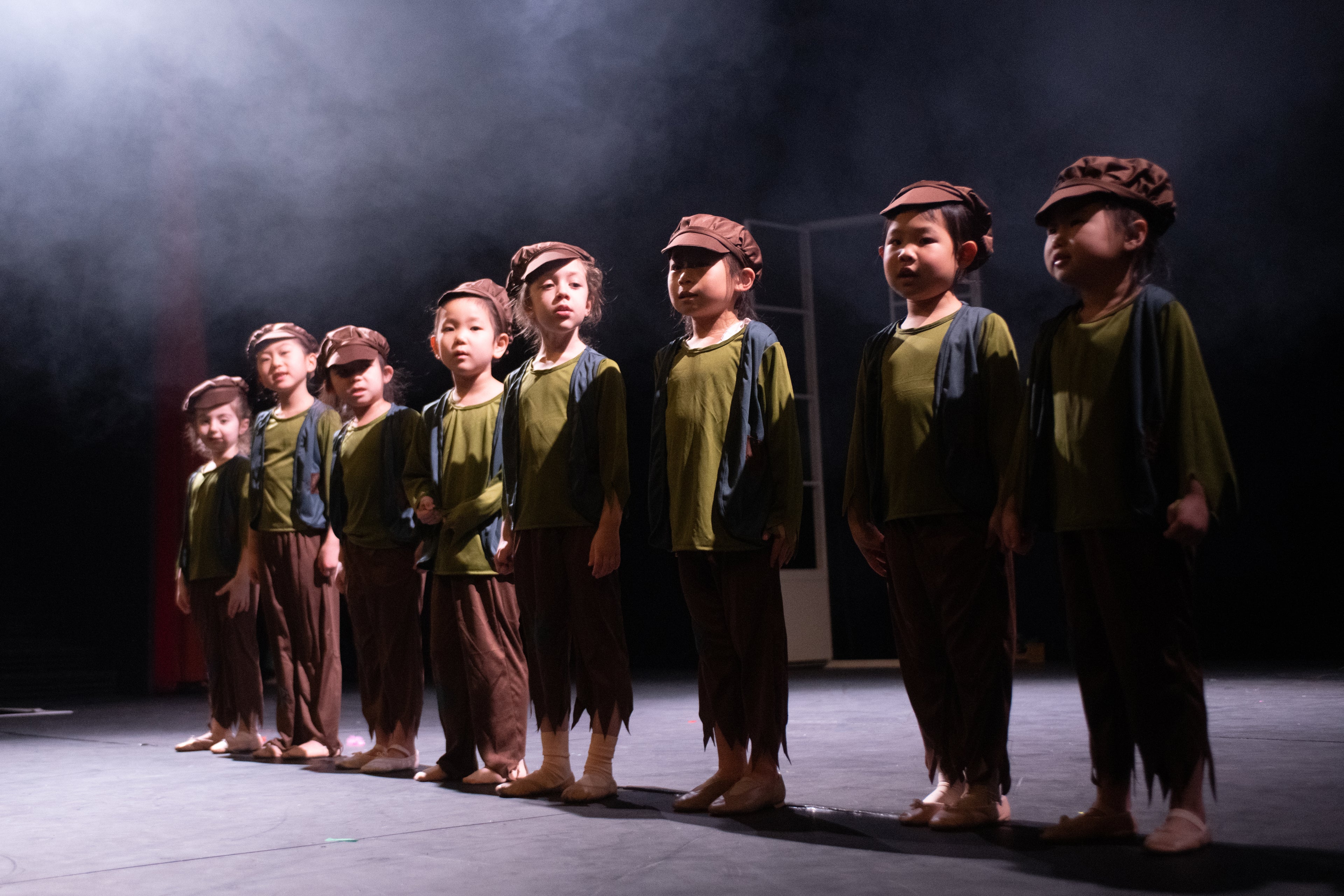 A line of young children on stage, dressed in green and brown costumes with hats, stand aligned under moody, smoky stage lighting, conveying focus and anticipation.
