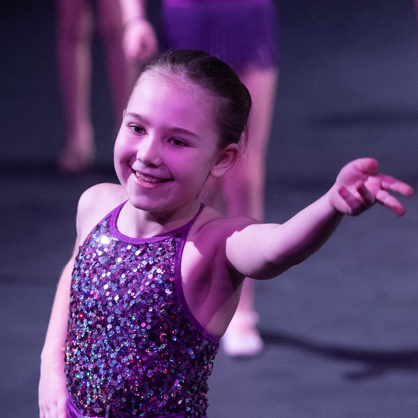 Smiling young girl in a sparkly purple dance costume performing on stage during a dance class.